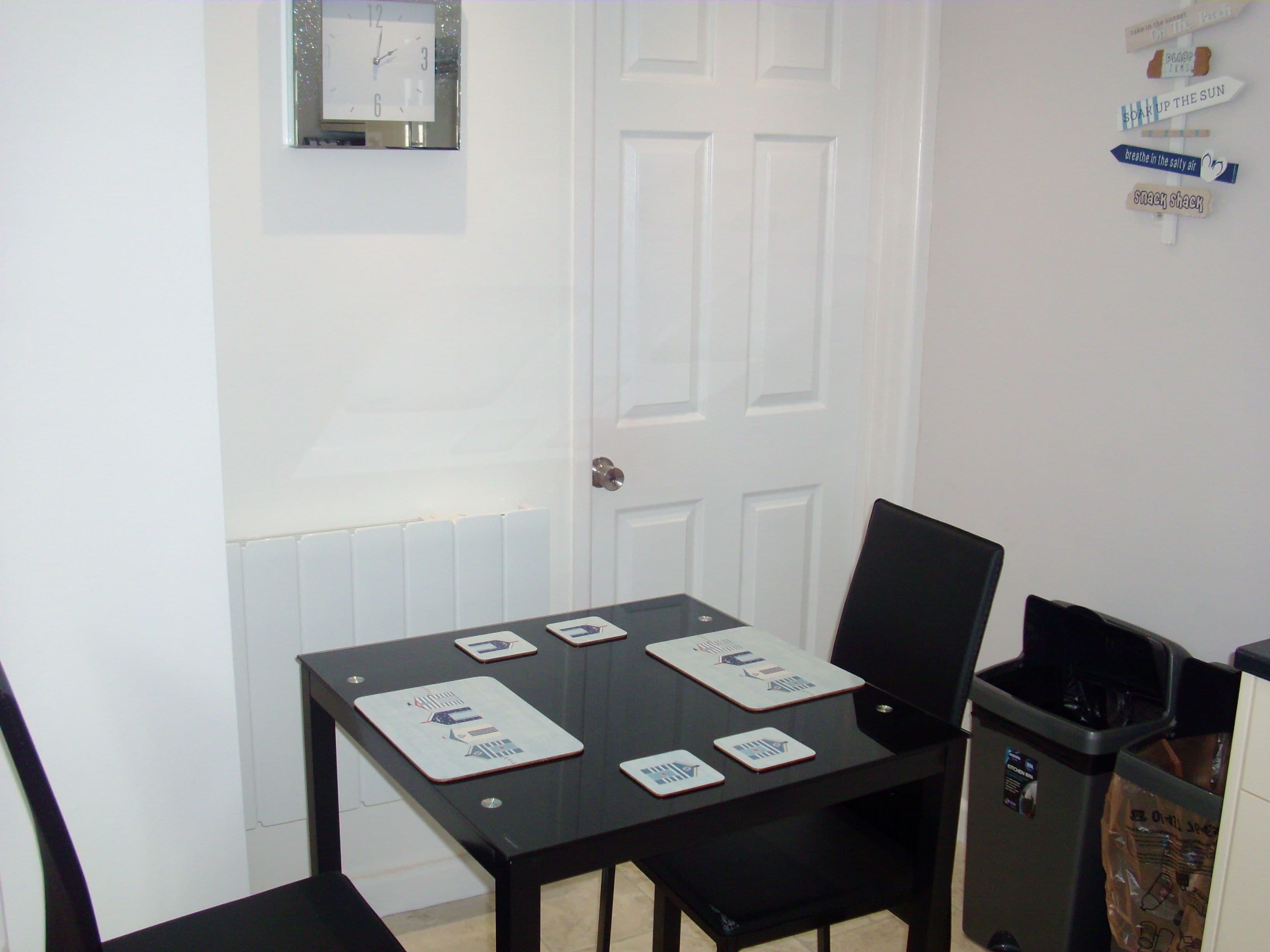 A small dining area with a table and chairs, a clock on the wall, and a trash bin.