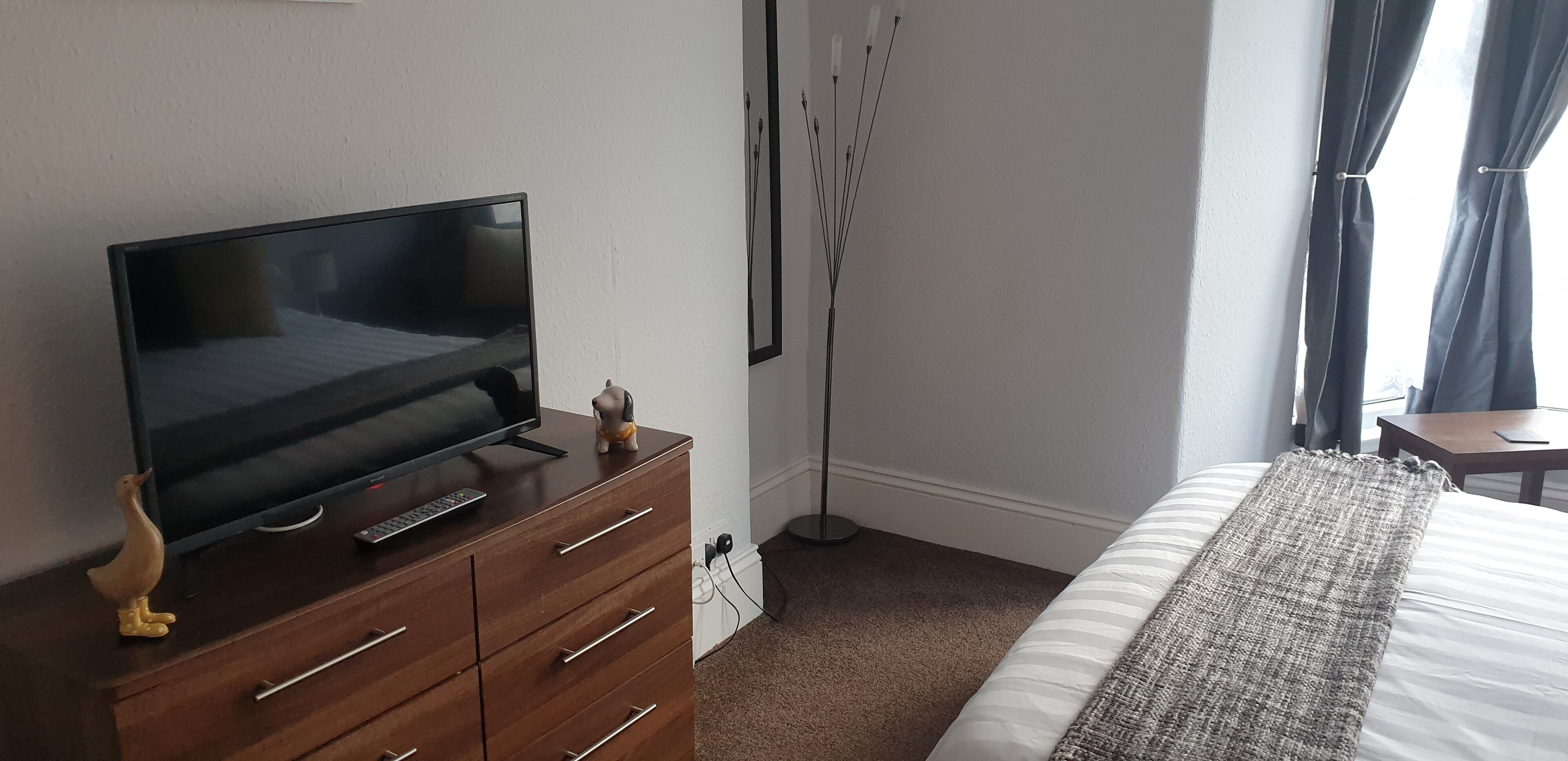 Habitación de hotel con televisor sobre un aparador de madera, una mesita de noche, una lámpara de pie y una cama con manta a rayas.