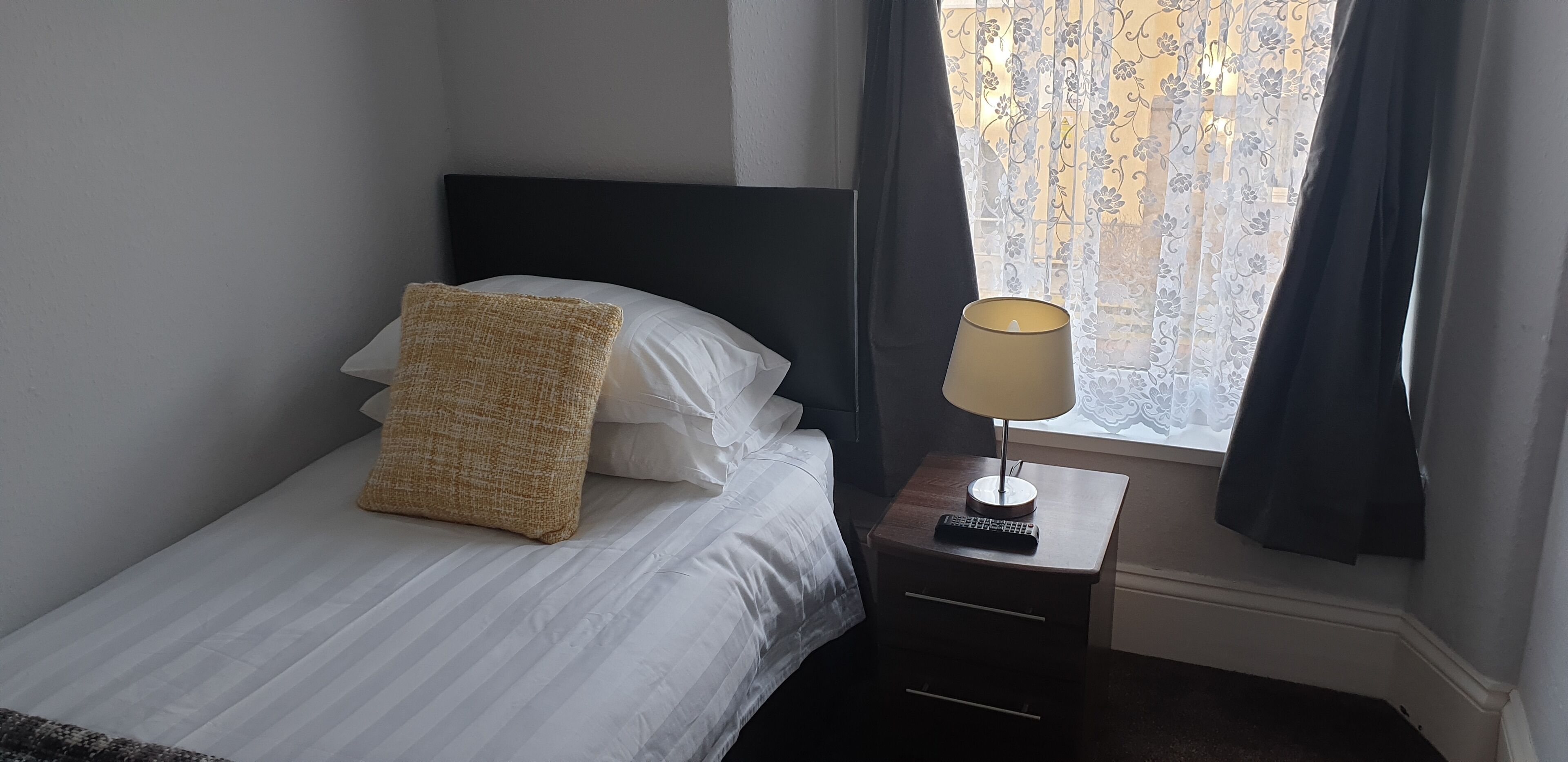 A neatly made bed with white linens and a textured throw blanket, a bedside lamp, and a remote control on a small wooden nightstand.