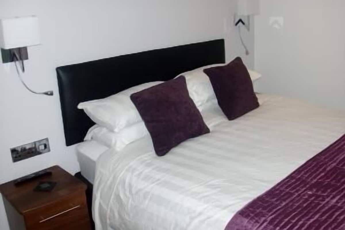 A neatly made bed with white and purple bedding, a black headboard, and a small wooden nightstand with a lamp.