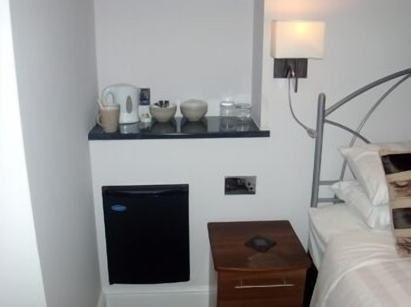 A hotel room with a bed, a bedside table, a small cabinet, a wall-mounted shelf with toiletries, and a wall lamp.