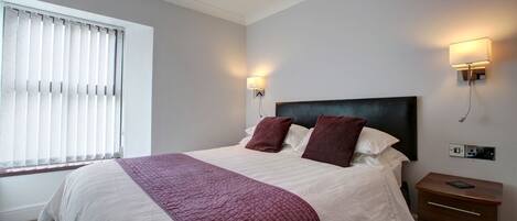 A bedroom with a bed, two bedside lamps, a nightstand, and a window with blinds.