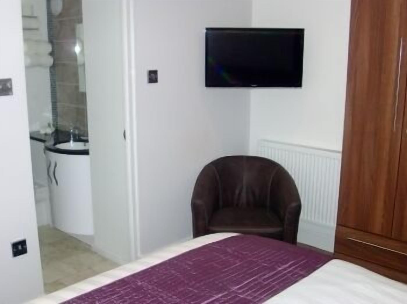 Un dormitorio con una cama, un televisor montado en la pared, una silla marrón y un armario de madera.