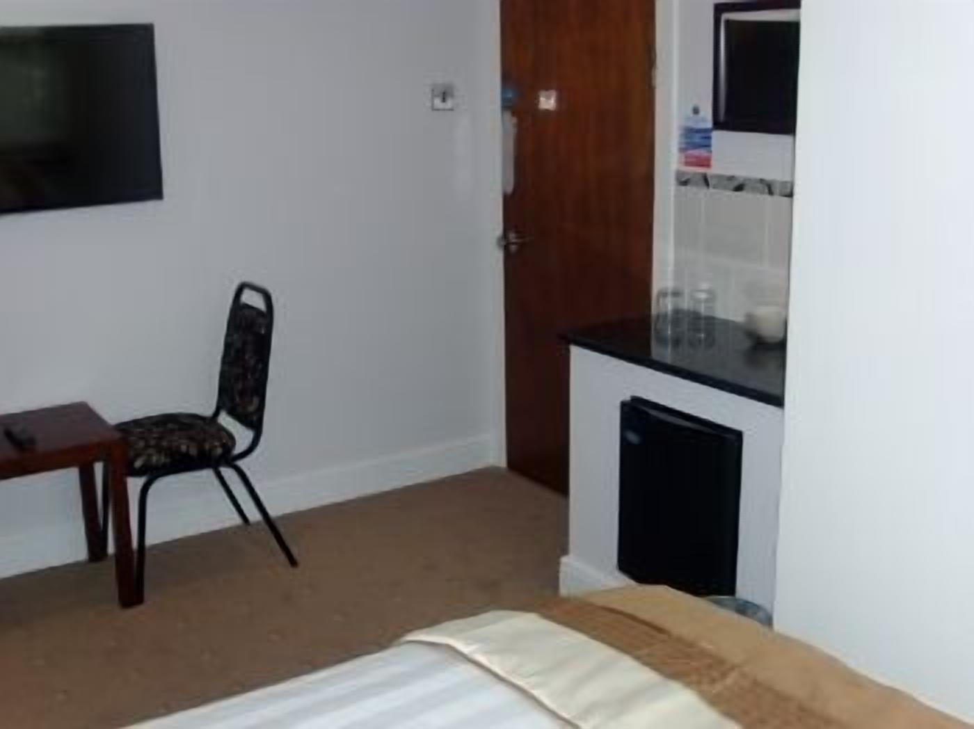 Une petite pièce comprenant un lit, un bureau, une chaise, un téléviseur fixé au mur et un coin cuisine avec un réfrigérateur et un four à micro-ondes.