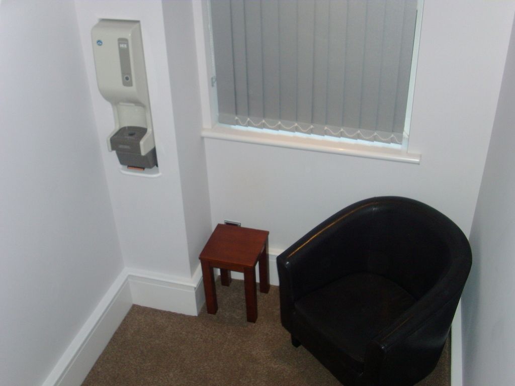 A corner room with a black leather chair, a small wooden table, and a white wall-mounted dispenser.