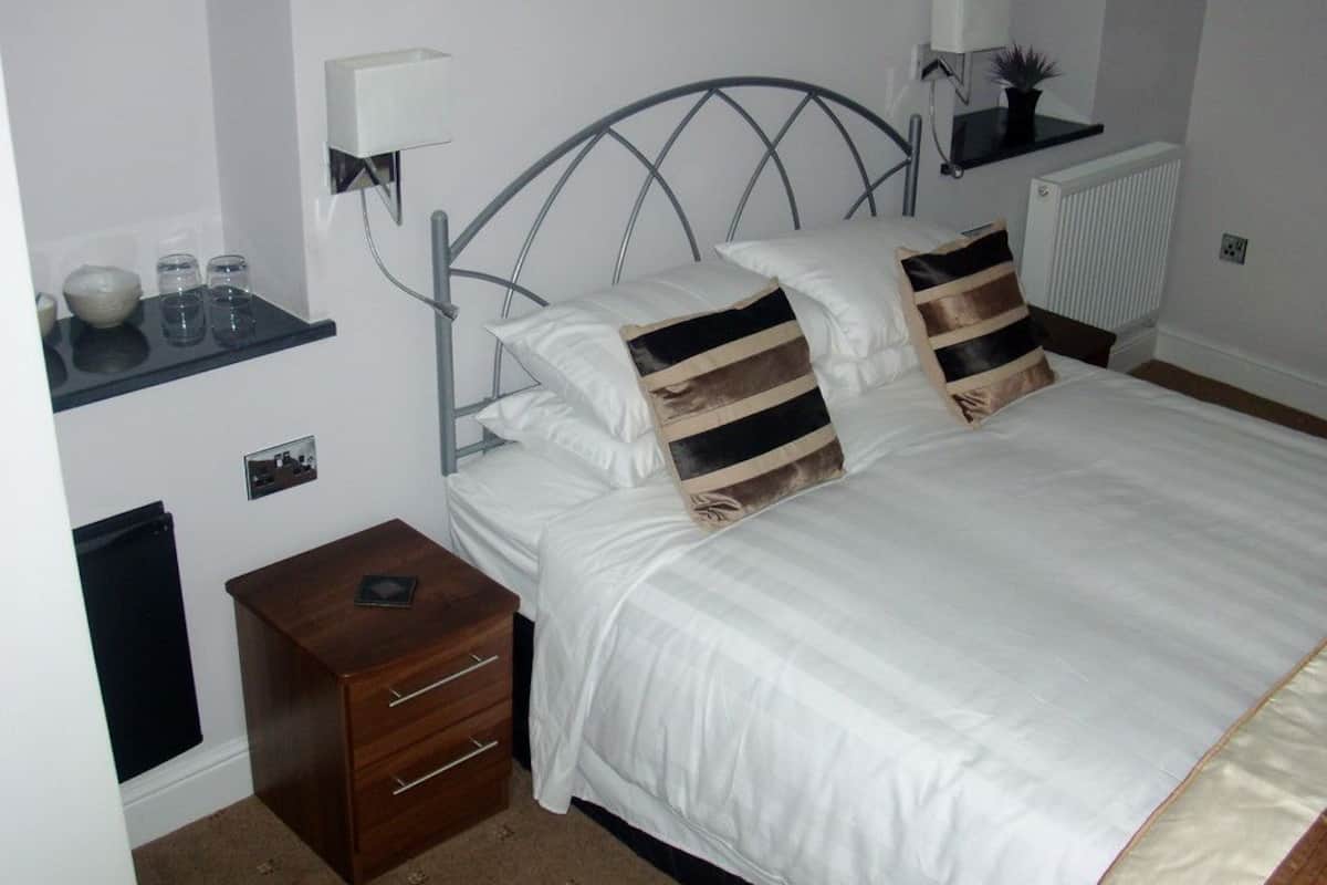 A neatly made bed with white bedding and striped pillows, a bedside table with a lamp, and a small shelf with glasses and a bowl.