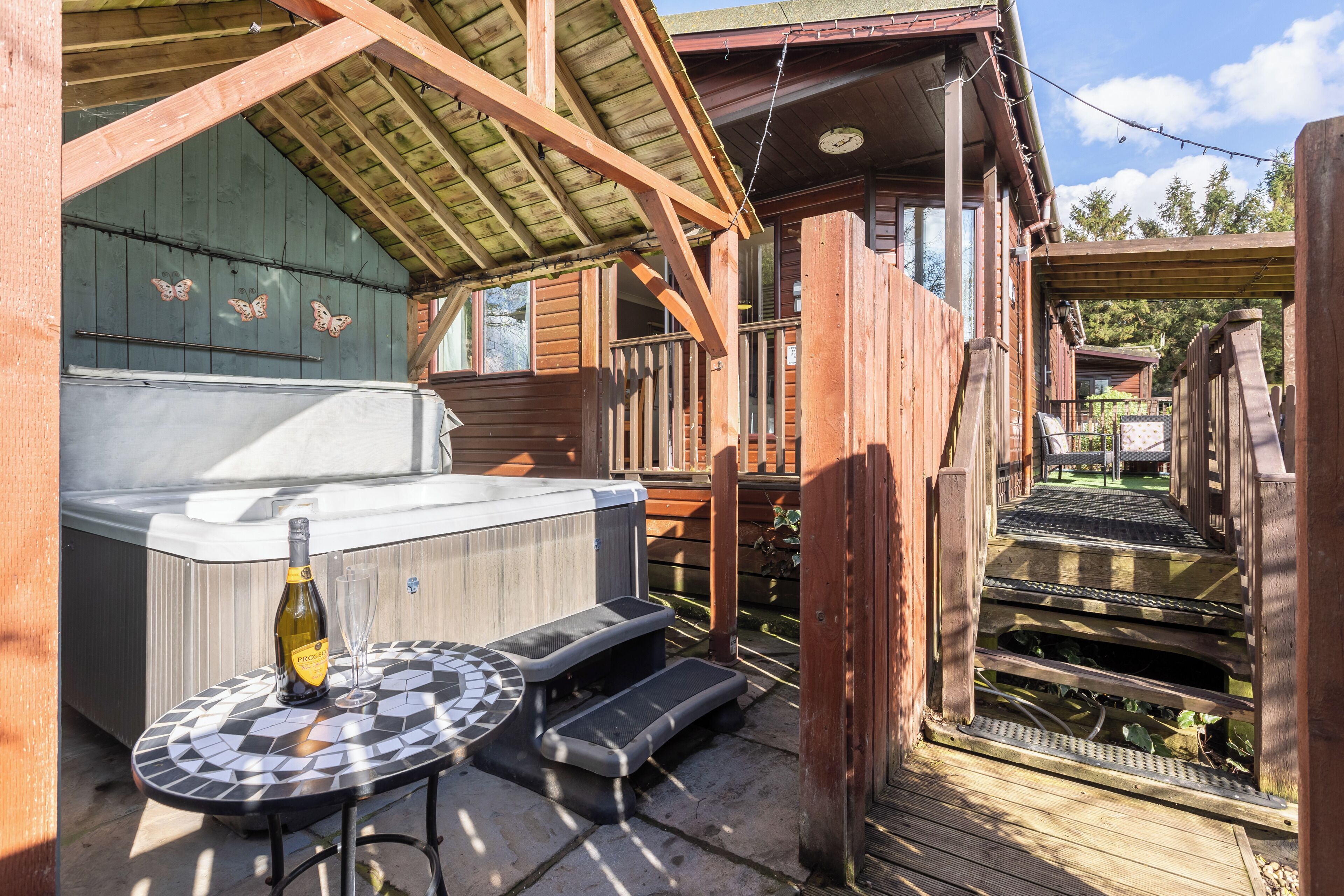 A wooden deck with a hot tub, a bottle of champagne, and a glass on a mosaic table.