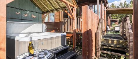 A wooden deck with a hot tub, a bottle of champagne, and a glass on a mosaic table.