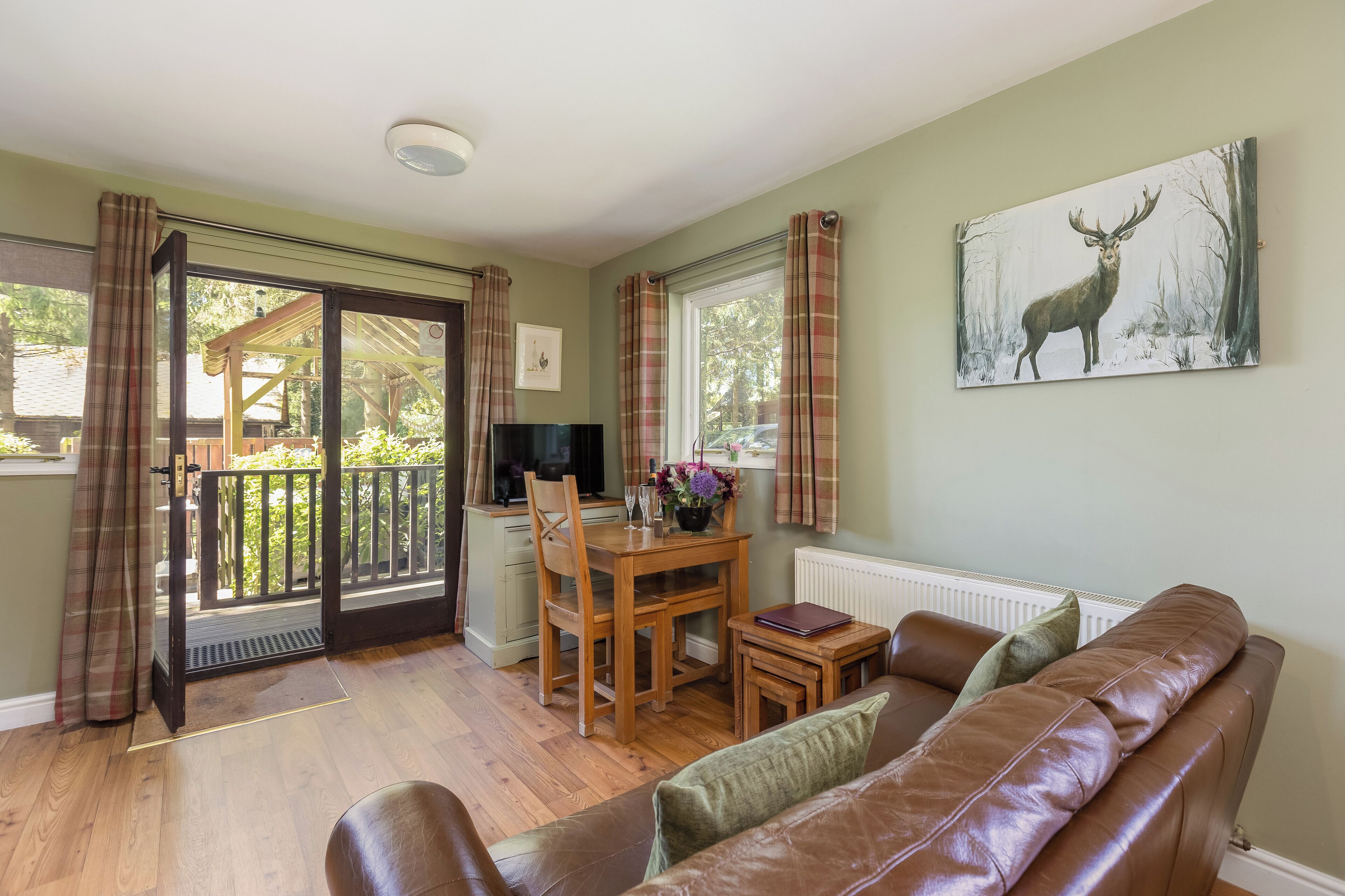 A cozy living room with a brown leather sofa, a wooden dining table with chairs, a television, and a painting of a deer on the wall.