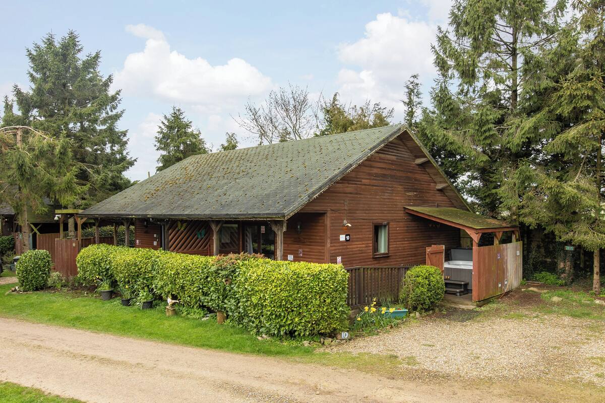 A rustic wooden cabin with a sloped roof, surrounded by dense greenery and tall trees.