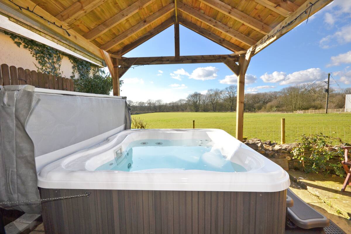 A hot tub under a wooden pergola with a view of a green field and trees.
