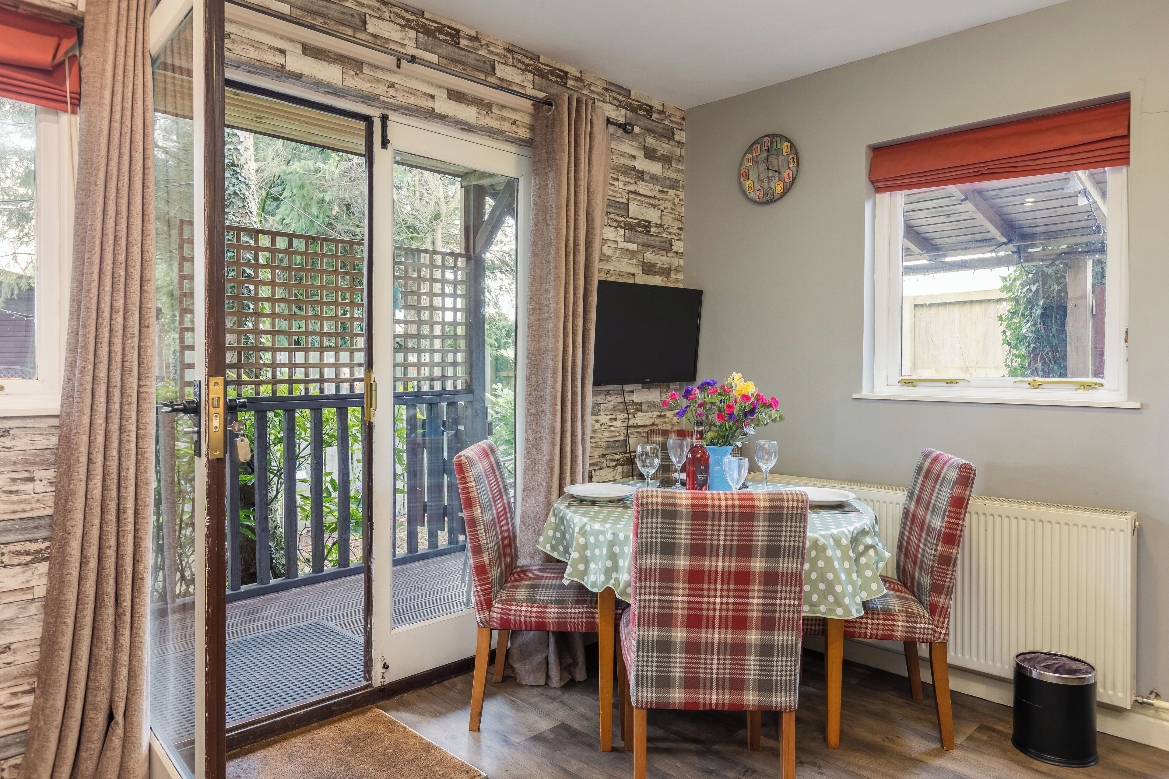 A dining area with a table set for two, plaid chairs, a TV mounted on the wall, and a window with a view of greenery.