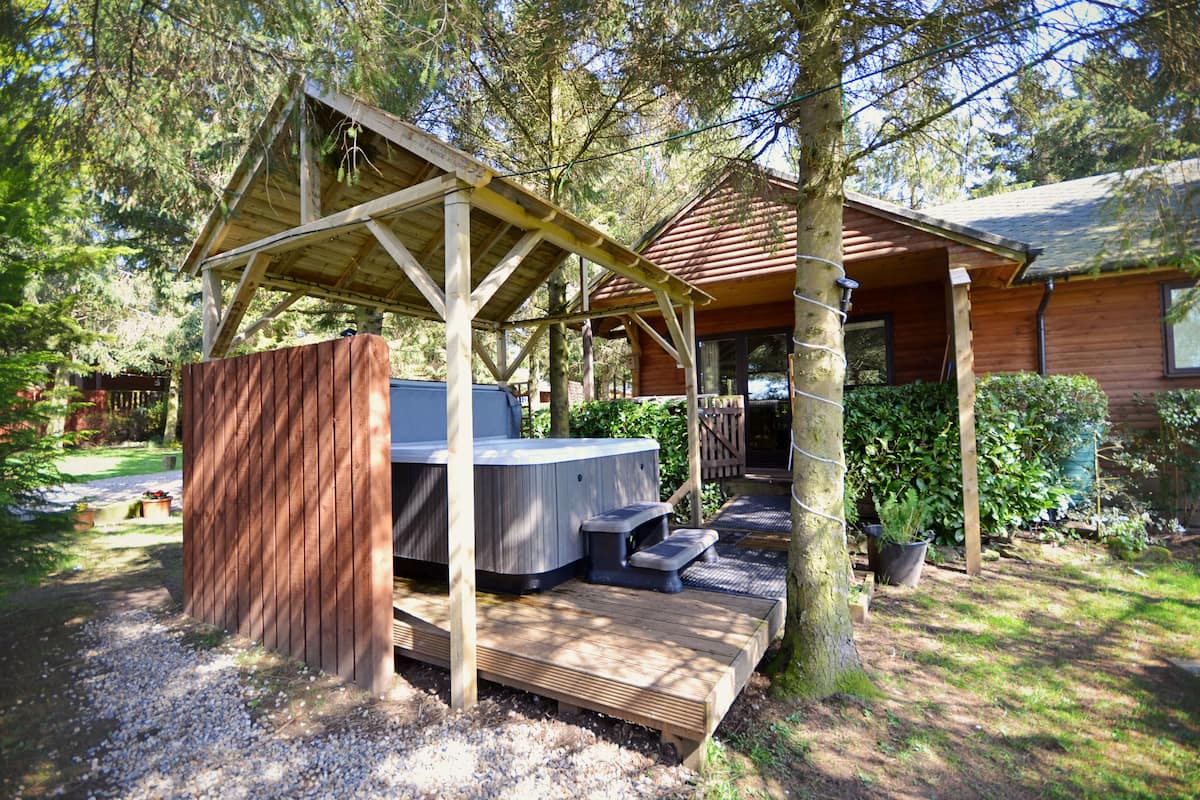 A wooden deck with a hot tub, a covered seating area, and a cabin in a wooded setting.