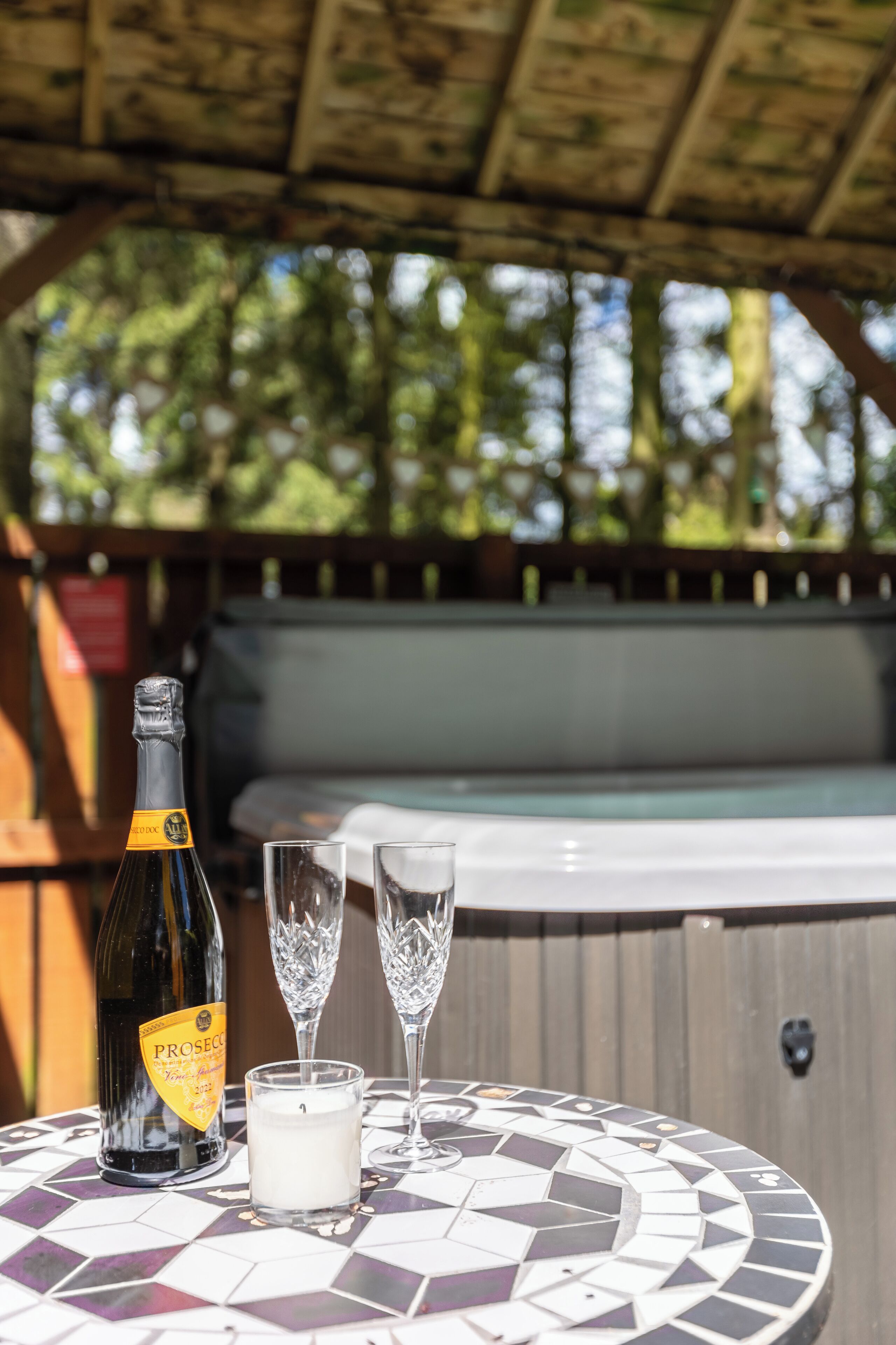 Een fles Prosecco, twee glazen en een kaars op een mozaiektafel onder een houten pergola.