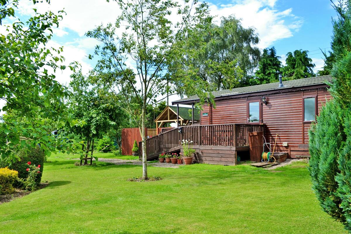 A wooden cabin with a deck, surrounded by a well-maintained lawn and various trees.