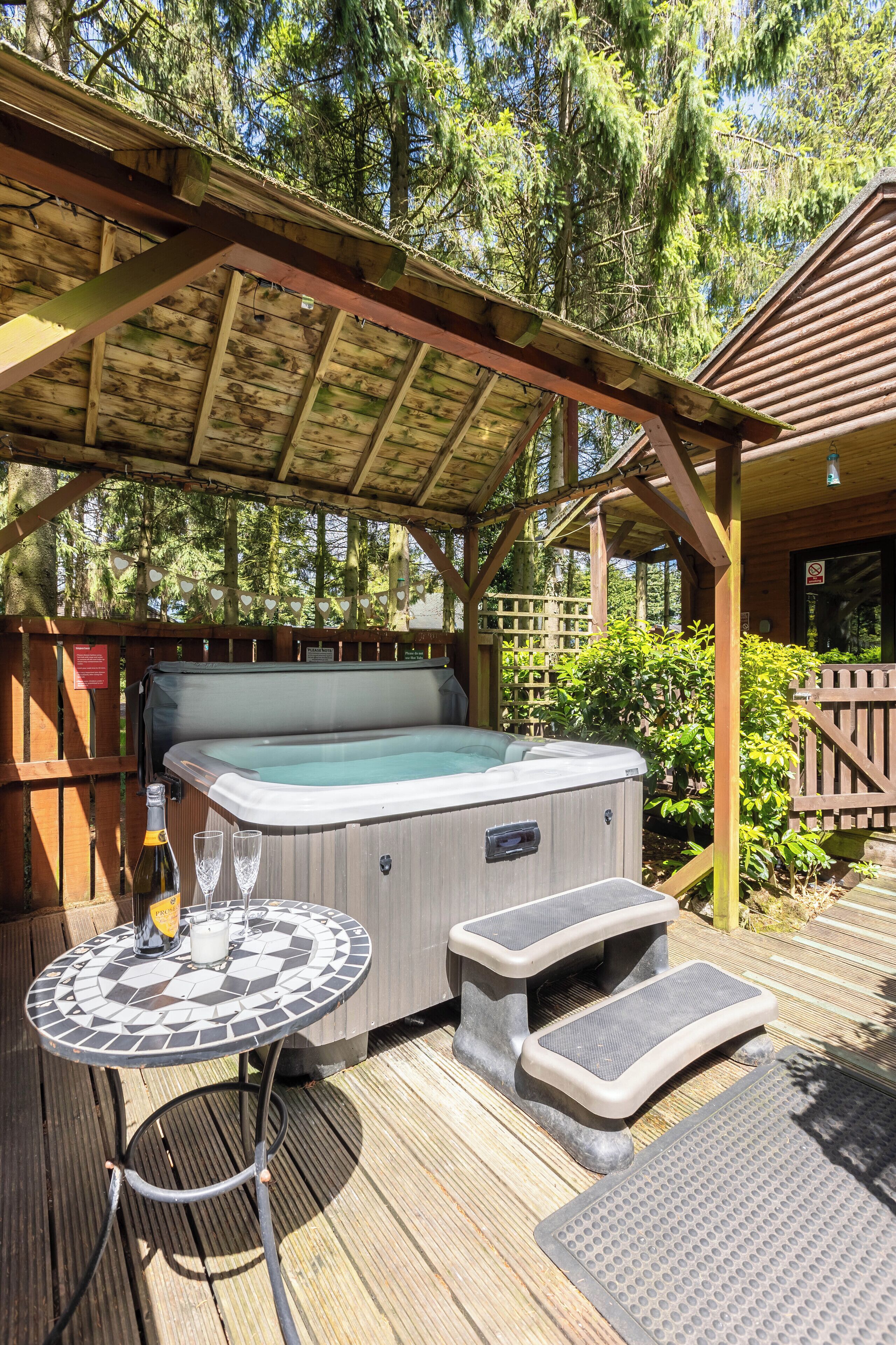 A hot tub with steps and a table with glasses and a bottle, surrounded by wooden decking and greenery.