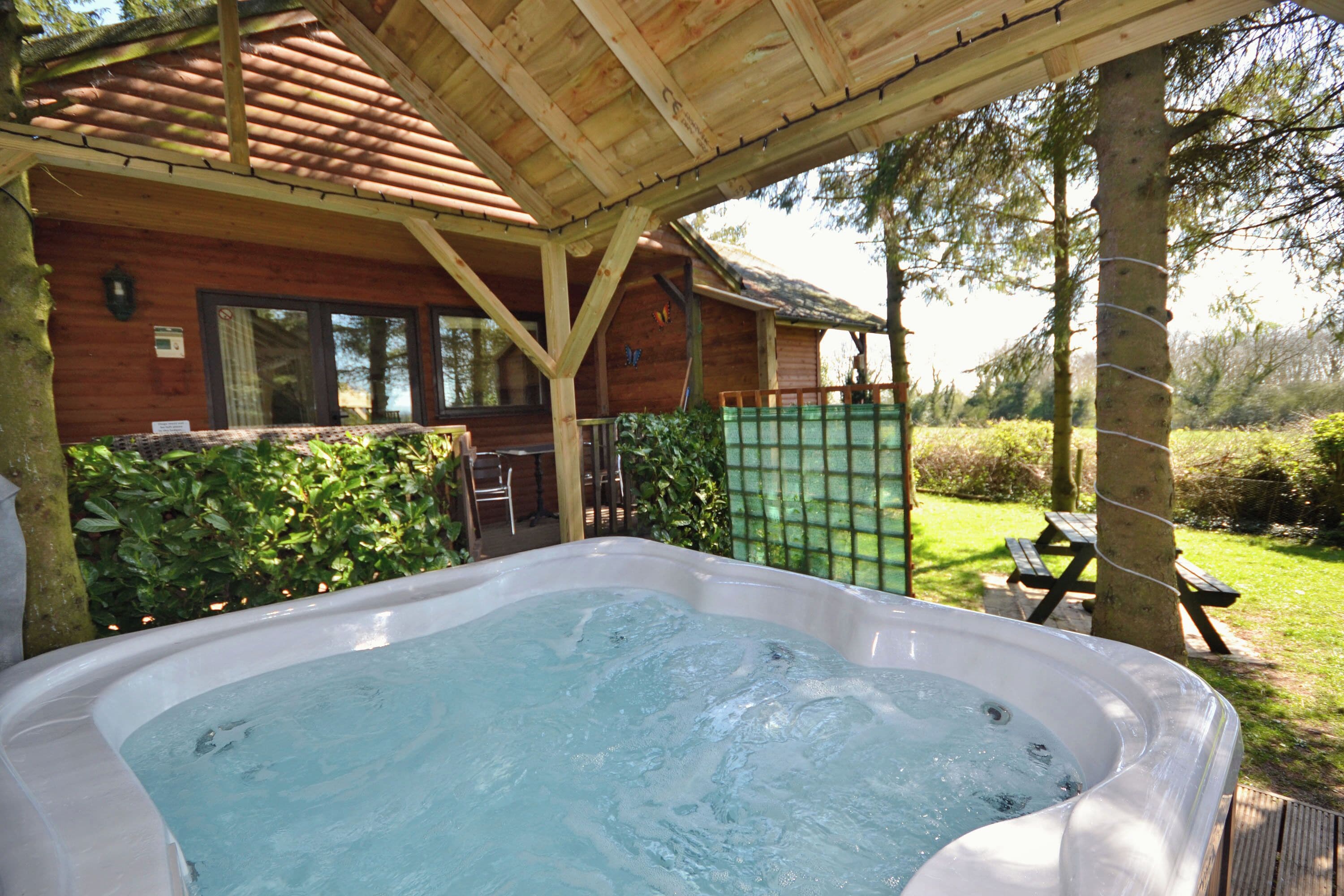 A hot tub in a covered outdoor area with a wooden deck, a picnic table, and a wooden cabin in the background.