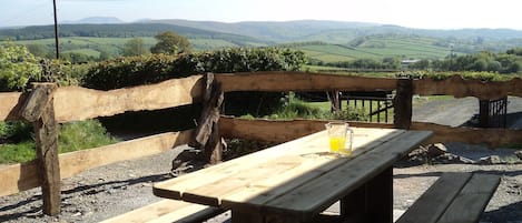 A wooden picnic table with benches, set on a gravel surface with a scenic view of rolling hills and fields in the background.