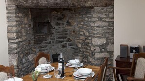 Una mesa de comedor preparada para cuatro, con platos blancos, copas de vino y una botella de vino. La mesa está frente a una pared de piedra con una chimenea.