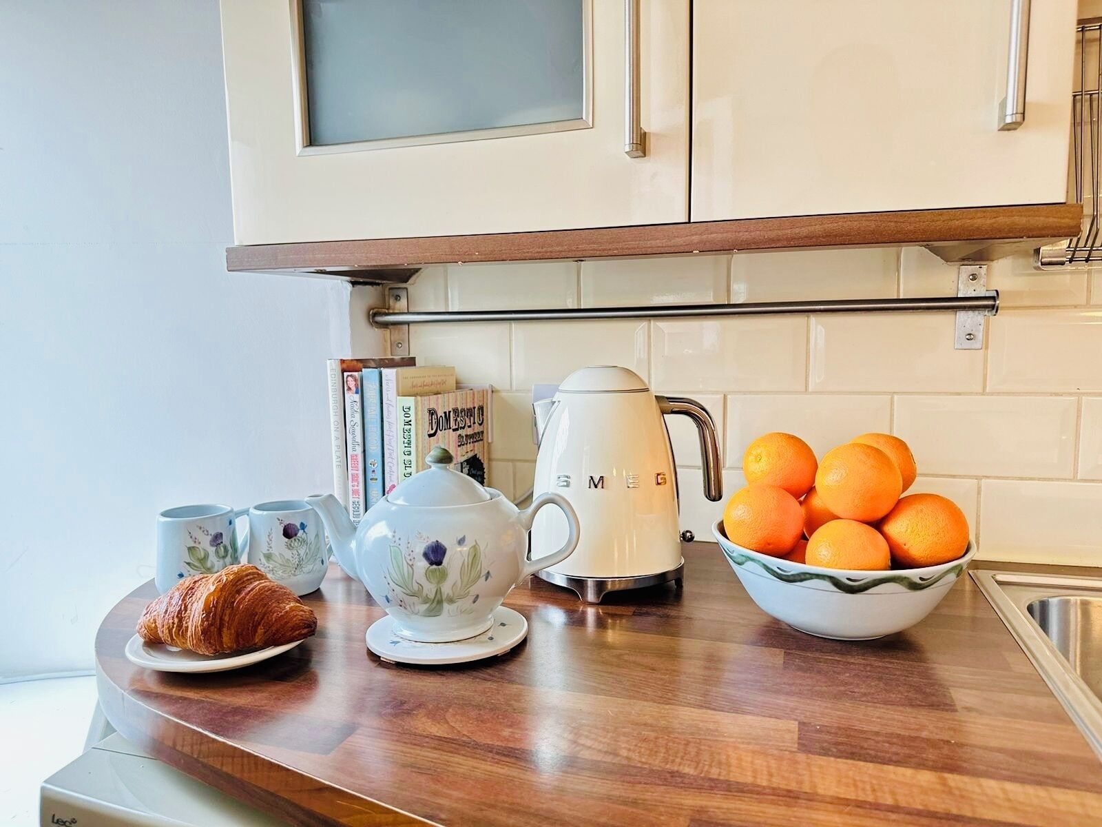 Een keukenblad met een koffiezetapparaat, theepot, croissant, boeken en een schaal mandarijnen.