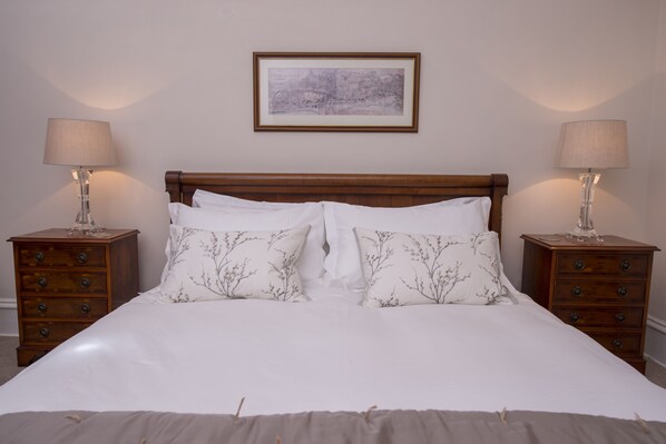 Una cama bien hecha con sábanas blancas y almohadas florales, flanqueada por dos mesitas de noche con lámparas, en una habitación con un cuadro en la pared.