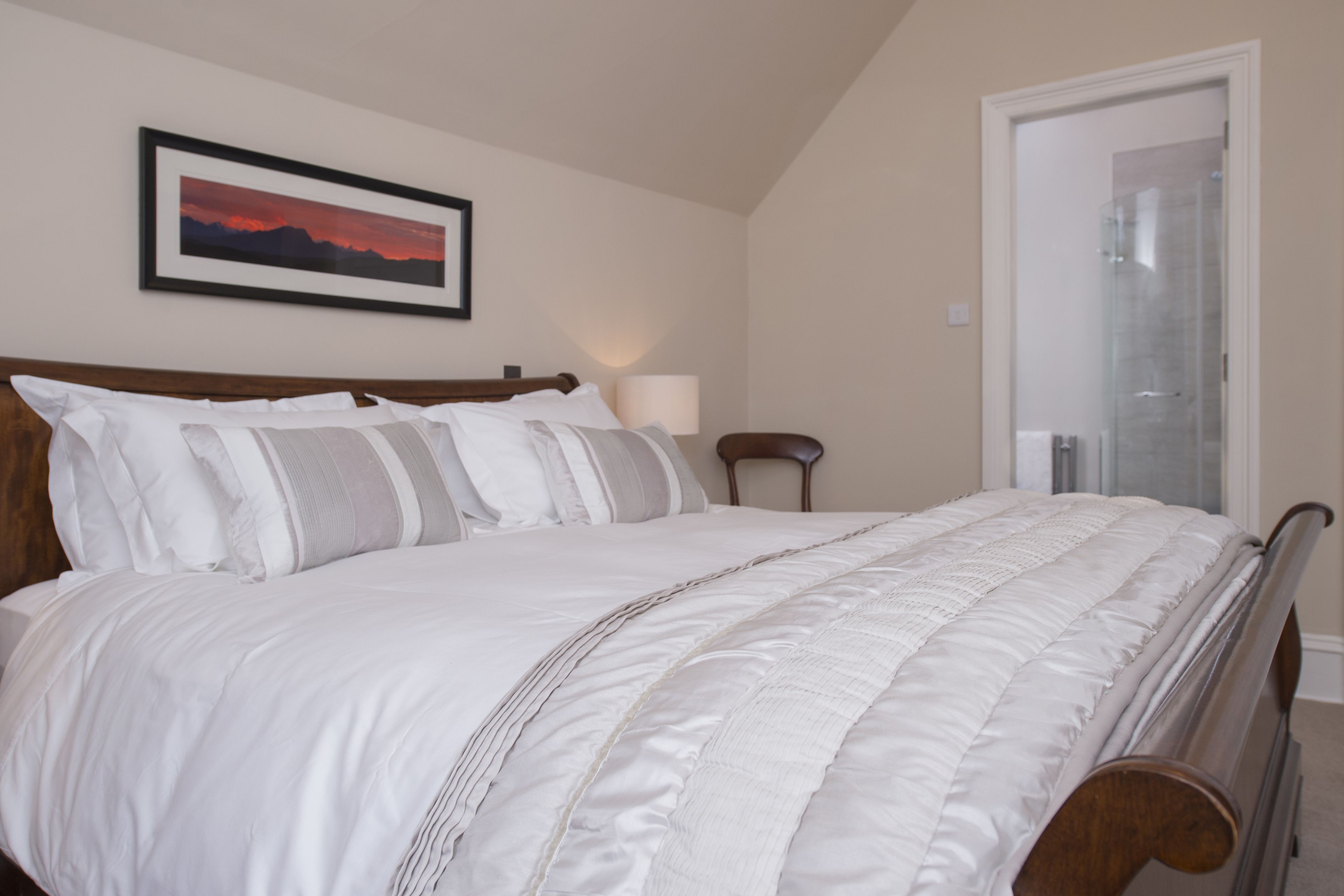 A neatly made bed with white bedding and a wooden headboard, a framed picture on the wall, and a door leading to a bathroom.