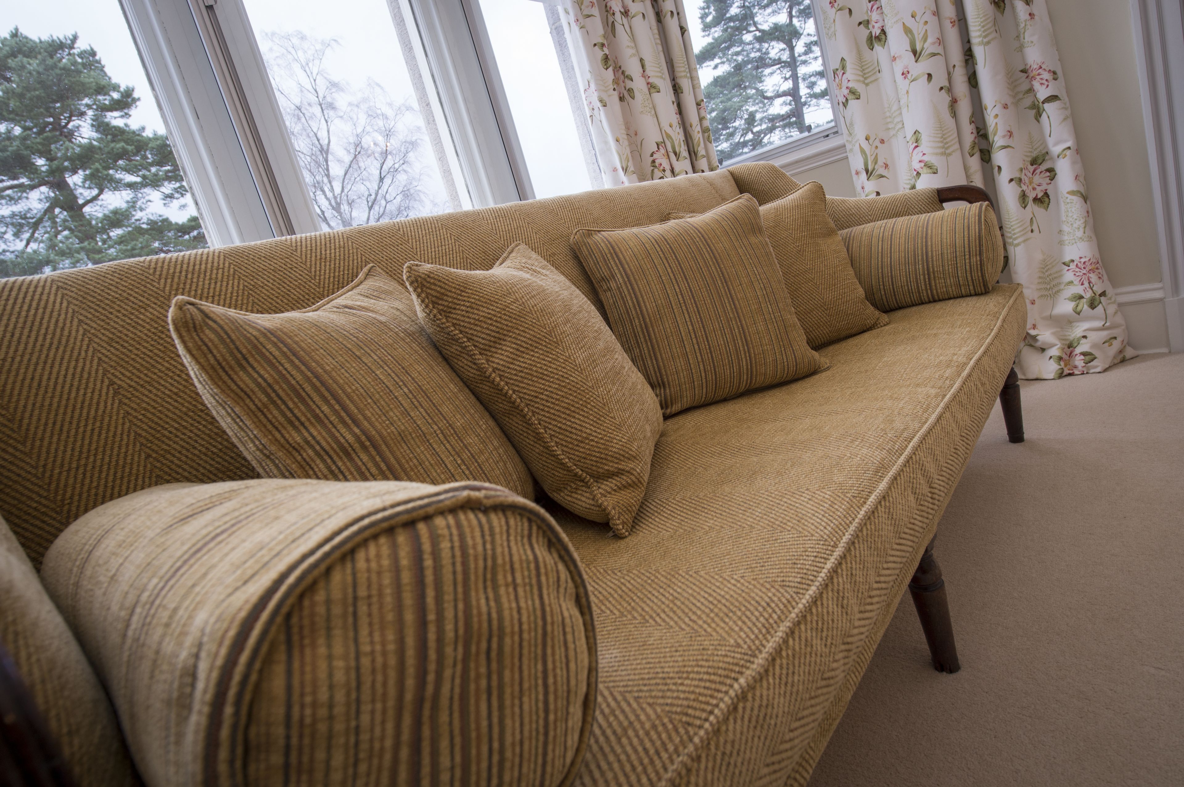 Ein beiges Sofa mit vielen Kissen in einem Raum mit großen Fenstern und blumigen Vorhängen.