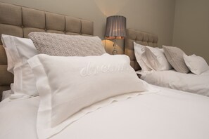 Una cama bien hecha con sábanas y almohadas blancas, un cabecero capitonado y una lámpara de noche.