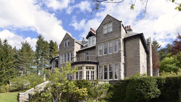 A large stone building with multiple windows and a curved balcony, surrounded by lush greenery and trees. - Lys-Na-Greyne Country House (Aboyne)