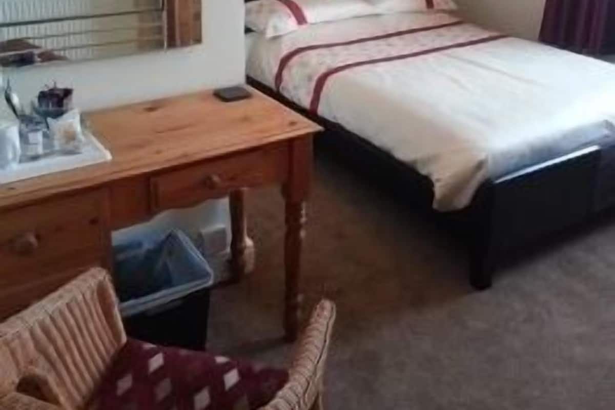 A hotel room with a bed, a wooden desk, a chair, a bedside table, a lamp, and a painting on the wall.