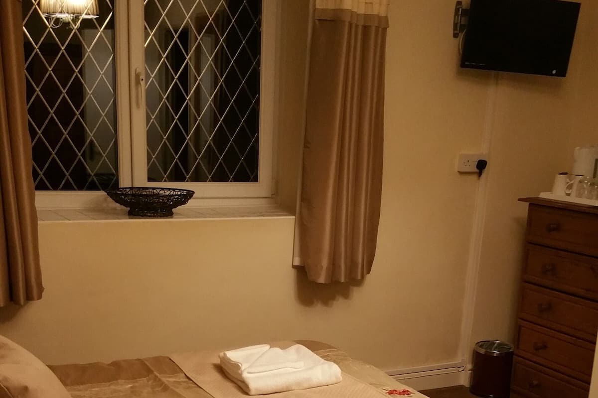 A neatly made bed with folded towels and a red booklet, a window with curtains, a television on the wall, and a wooden dresser with a trash can beside it.