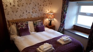 Un lit bien fait, avec une literie blanche et violette, un mur orné d’un motif floral et une fenêtre avec des rideaux.
