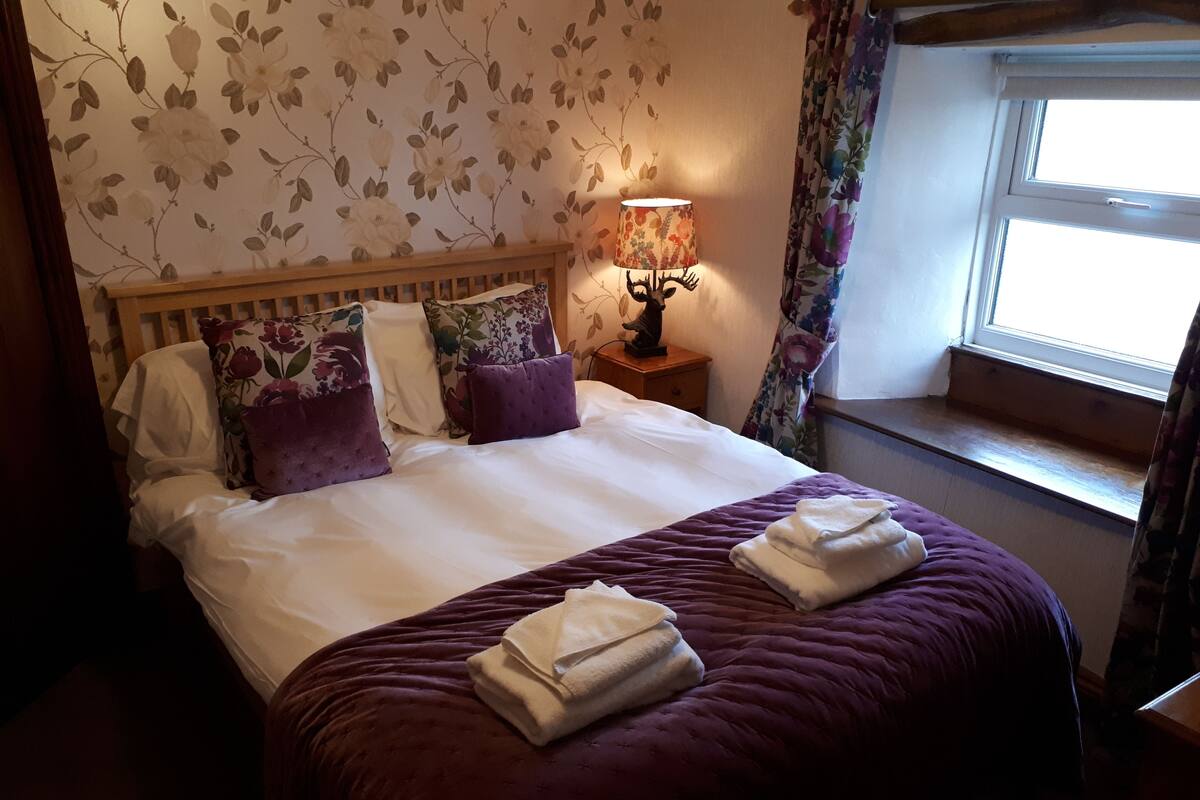 Ein ordentlich bezogenes Bett mit weißer und violetter Bettwäsche, eine Wand mit floralem Muster und ein Fenster mit Vorhängen.