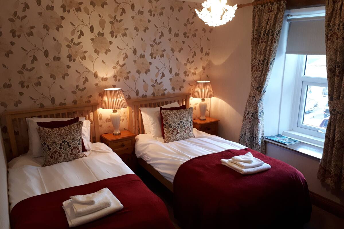 Ein Doppelzimmer mit zwei Betten, jeweils mit roter Tagesdecke und weißen Laken. Zwei Nachttische mit Lampen und ein Fenster mit Vorhängen.