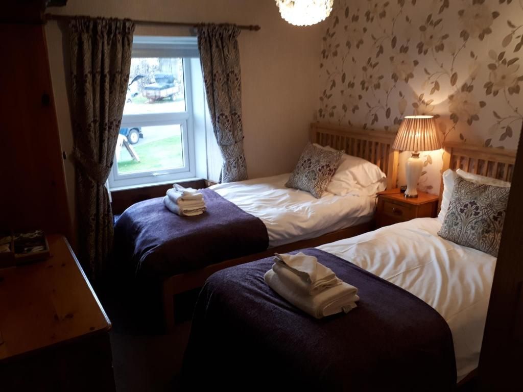 A room with two beds, a window with curtains, a lamp on the nightstand, and a wooden table.