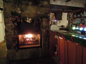 Une cheminée en pierre avec un feu allumé, entourée de décorations festives. Un coin bar avec un comptoir et des étagères remplies de bouteilles est visible sur la droite.