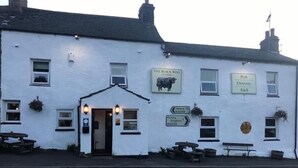 Un bâtiment blanc avec des enseignes indiquant le pub « The Black Bull » et l’établissement proposant à la fois un restaurant et des chambres d’hôtes. On y trouve également des panneaux de signalisation indiquant la direction de Hawes et de Kirkby Stephen.