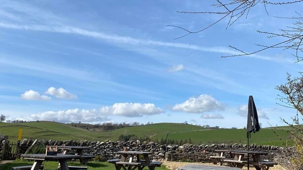 Un espace pique-nique doté de nombreuses tables et bancs en bois, entouré d’un mur de pierre et de prairies verdoyantes, le tout sous un ciel dégagé.