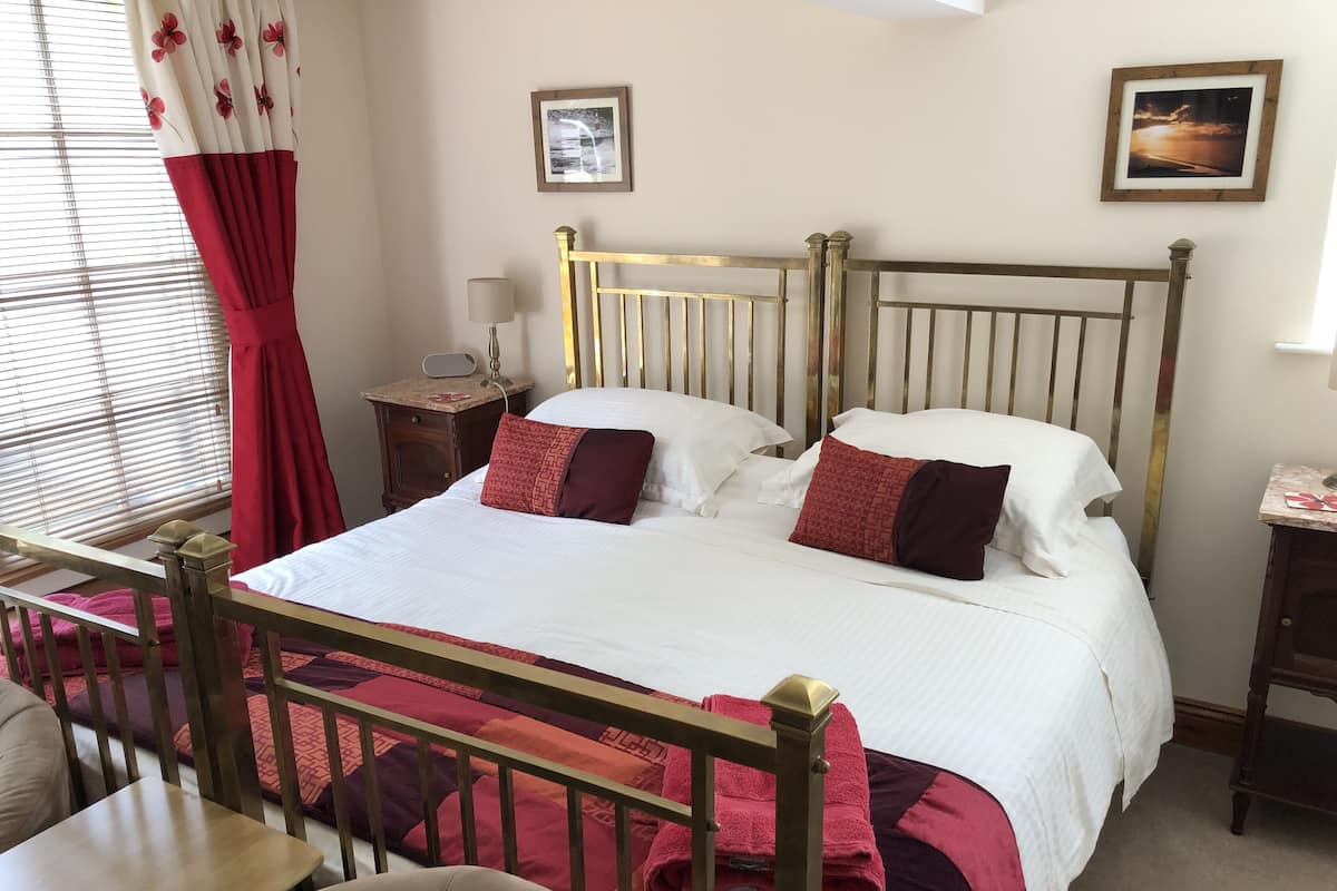 A neatly made bed with a wooden headboard, a bedside table with a lamp, and a window with blinds and red curtains.