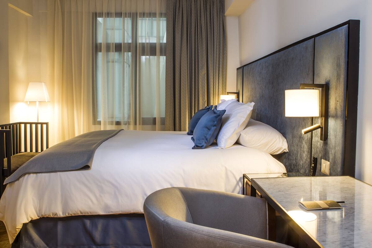 A hotel room with a large bed, a desk with a lamp, and a view of the outside through sheer curtains.