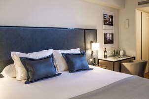 Habitación de hotel con una cama grande, dos almohadas, una mesita de noche con una lámpara, una silla y una pequeña mesa con una botella y vasos.