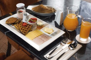 Bandeja de desayuno con waffles, tostadas, fruta, queso y una jarra de jugo de naranja sobre una mesa de mármol.