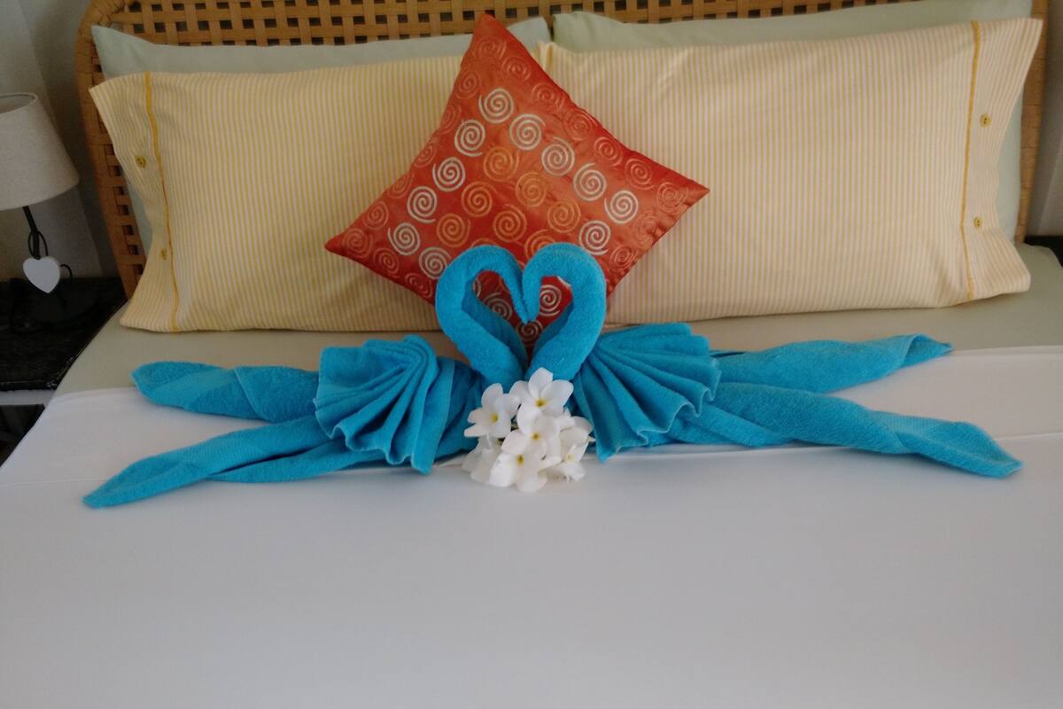 A bed with a white sheet, a blue towel arranged in a heart shape, a white flower, and a red pillow with a swirl pattern.