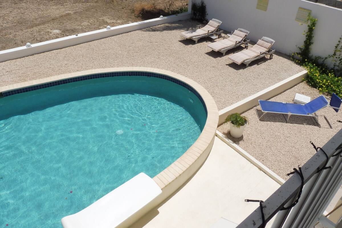 A swimming pool with a curved edge, surrounded by a gravel area with lounge chairs and umbrellas.
