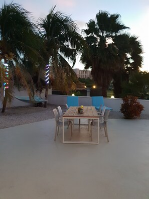 An outdoor patio with a wooden table and chairs, surrounded by palm trees and a hammock. - Karibu Aruba Boutique Hotel (Noord)