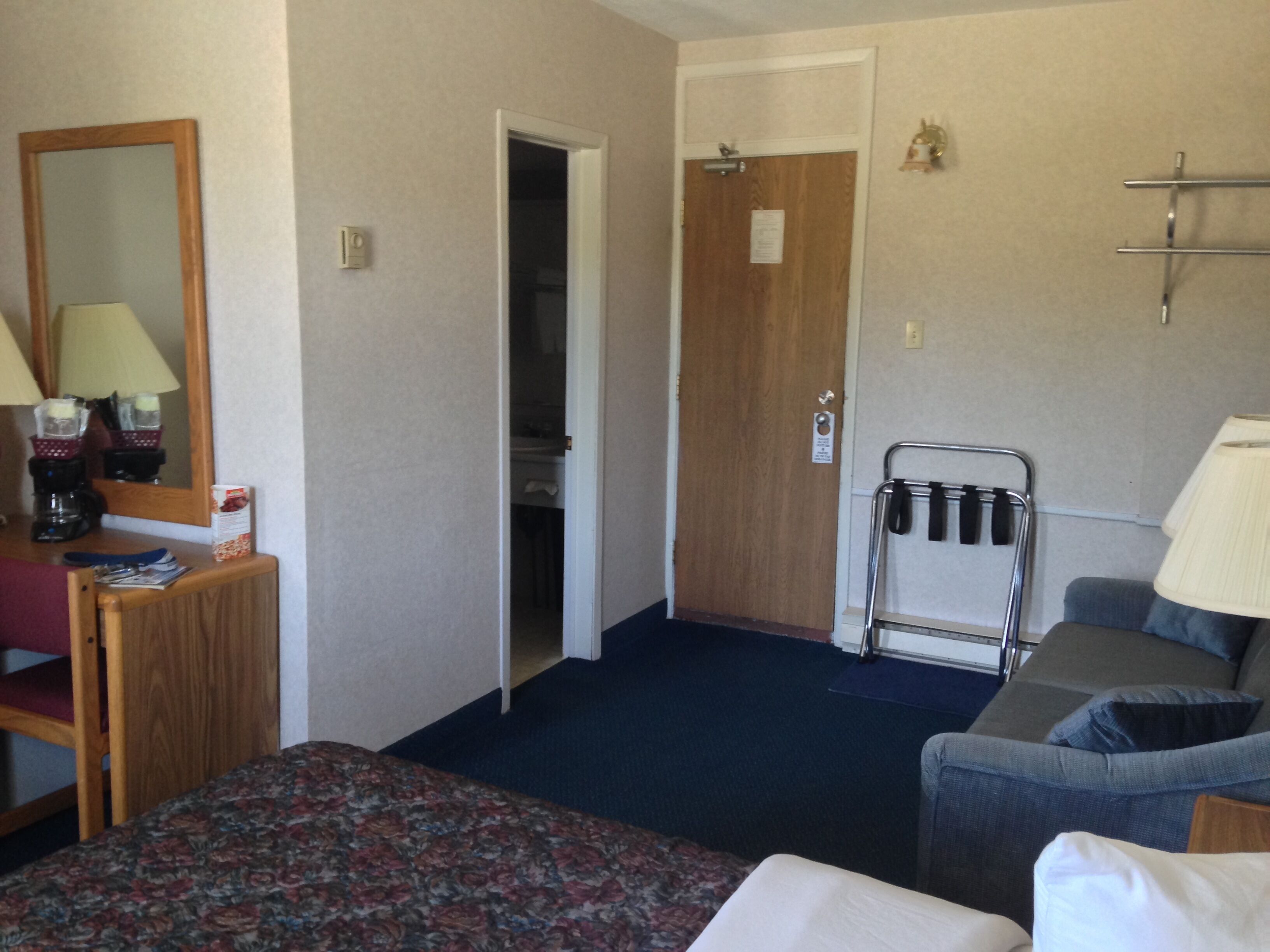 Een hotelkamer met een bed, een bureau met lamp, een stoel, een kapstok en een deur.