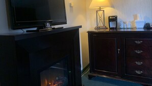 A flat-screen TV on a stand above a fireplace, a lamp on a table, and a wooden cabinet with drawers.
