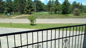 A view from a balcony with a metal railing, overlooking a road and a green landscape with trees and a small bush.