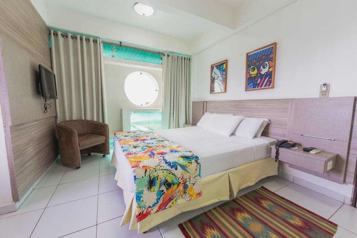 A hotel room with a bed, a chair, a desk, a TV, and artwork on the walls.