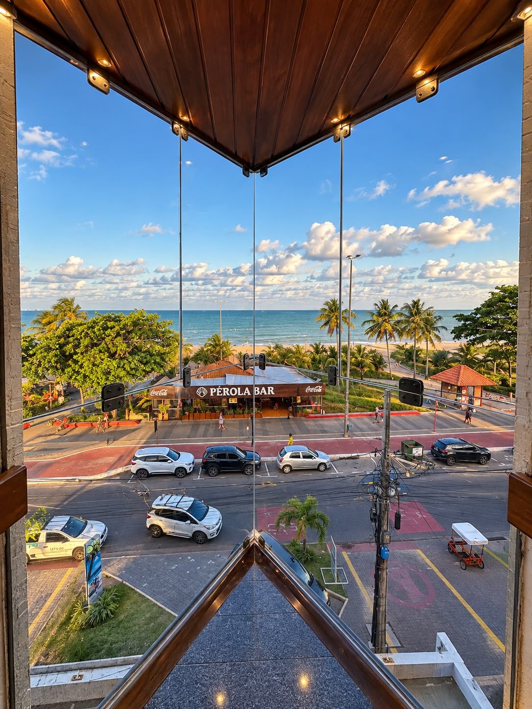 Vista da janela com rua movimentada, um bar chamado 'Perola Bar' e uma praia ao longe.